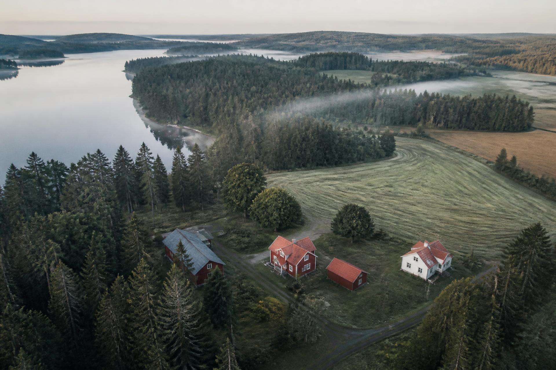 Flygbild över skog, sjö och gårdsbyggnader i morgondimma